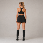Back view of the black Fitfigura Club Skirt – Luxe Resort Form, showing the rear fit of the skirt styled with a matching black sports bra and tall boots.