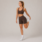 Model wearing the dark brown Fitfigura Contour Bra – Defined Sculpt in a standing leg-lift pose, highlighting the flexible performance fit.