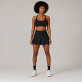 Model wearing the Fitfigura Court Skirt – Classic Elite Form in black with a matching black sports bra, black socks, and white sneakers, shown from the front.