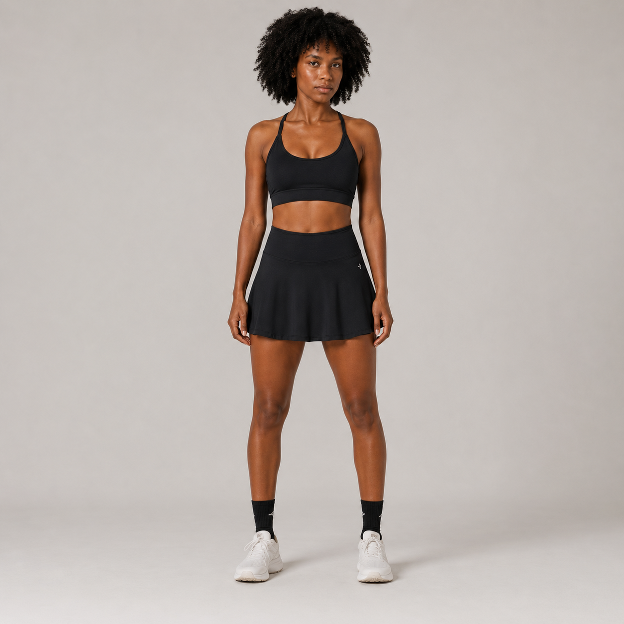 Model wearing the Fitfigura Court Skirt – Classic Elite Form in black with a matching black sports bra, black socks, and white sneakers, shown from the front.