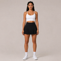 Model wearing the Fitfigura Rally Tennis Skirt – Sculpt Court Form in black with a white crop top, white socks, and white sneakers, shown from the front.