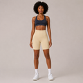 Model wearing the Fitfigura Studio Short 8” – Flow Essential Form in cream with a blue sports bra and white socks, shown from the front.