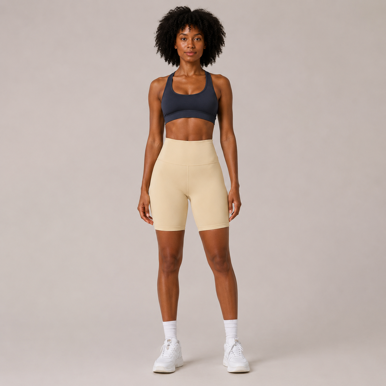 Model wearing the Fitfigura Studio Short 8” – Flow Essential Form in cream with a blue sports bra and white socks, shown from the front.